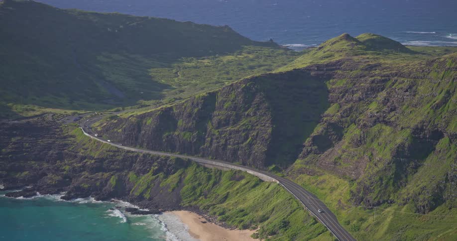 A stunning aerial view shows cars driving along a scenic coastal highway that winds through the lush green mountains of a tropical island