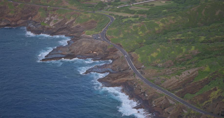 An epic aerial view of cars traveling along a scenic coastal highway carved into the side of a lush, volcanic island as ocean waves crash against the rocky shore