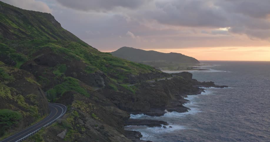 Aerial view slowly rises over a scenic coastal highway nestled between green mountains and the ocean at sunset