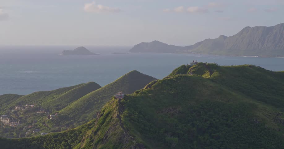 A majestic zoom-out from a verdant mountain ridge reveals hikers enjoying the panoramic ocean view and the expansive Hawaiian coastline