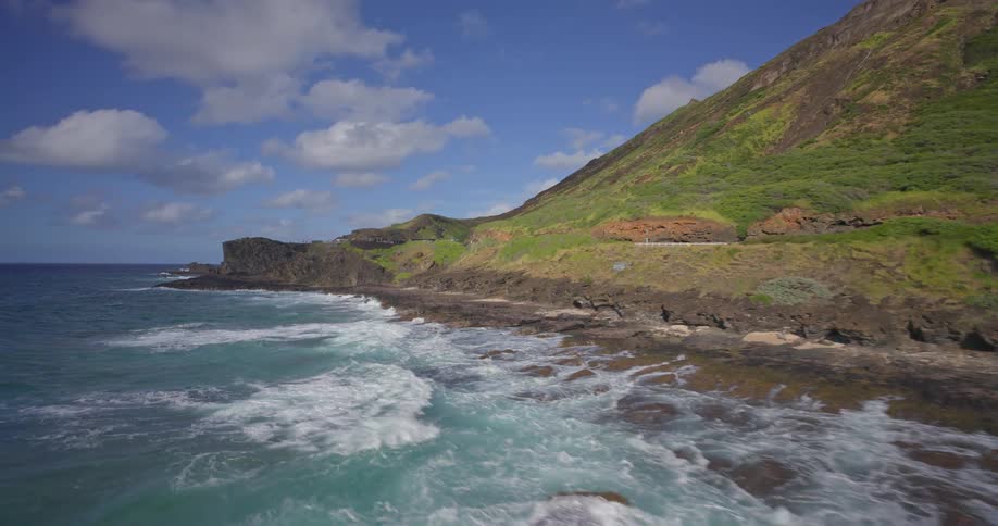 A stunning aerial view flies over a sandy beach and along the rugged volcanic coastline of a lush green Hawaiian island