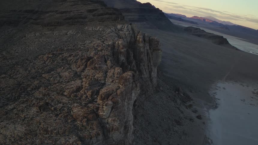 An aerial view reveals the vast, rocky landscape of a desert mountain range bordering a dry salt flat at sunrise