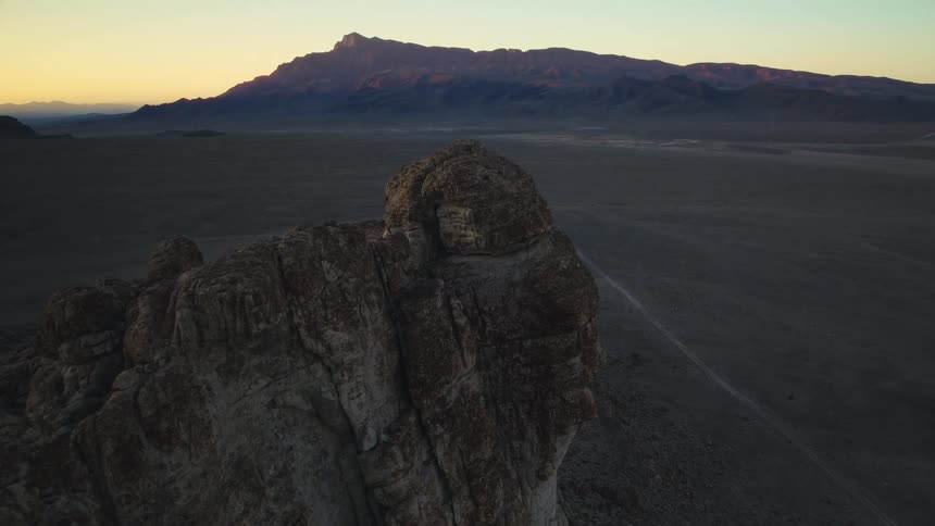 A cinematic aerial view slowly approaches a rugged cliff in a vast desert landscape as the sun rises over a distant mountain range