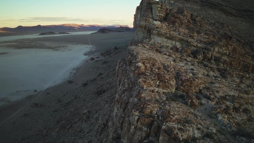 A slow cinematic aerial shot flies along the edge of a massive, rocky cliff overlooking a vast desert landscape at sunrise