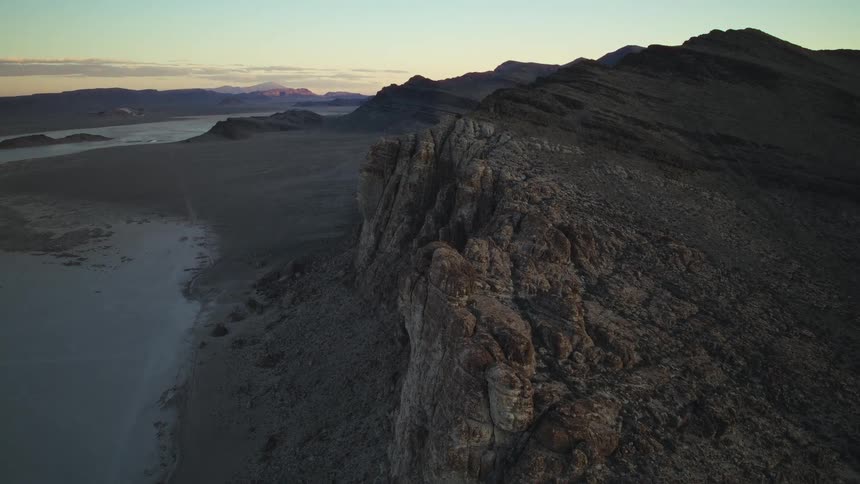 An aerial view shows a rugged mountain ridge overlooking a vast desert plain during a serene sunset