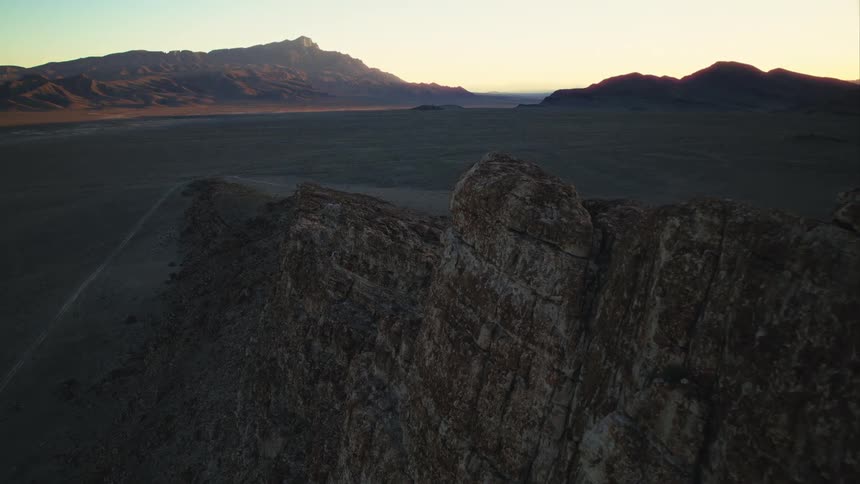 Drone footage slowly reveals a majestic desert valley and distant mountains from behind a rocky ridge at sunrise
