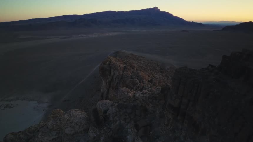 An aerial view slowly advances along a rugged mountain crest above a wide desert valley during twilight