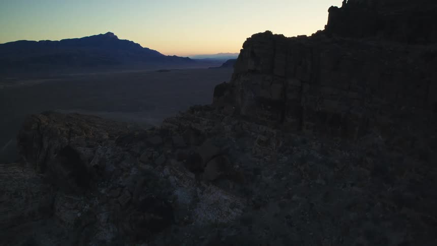 A slow aerial push over a dark, rocky mountain ridge reveals a vast desert valley at twilight