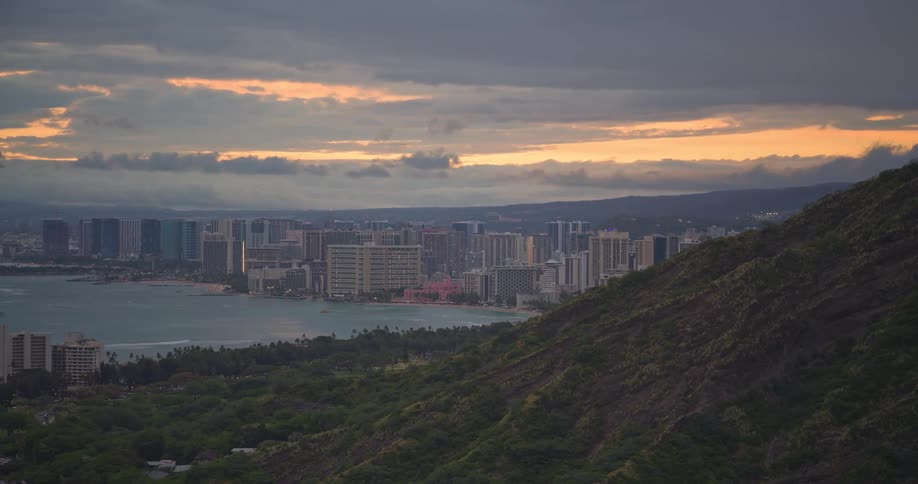 A stunning view of the Honolulu city skyline and Waikiki Beach from a mountainside at sunset under a cloudy sky
