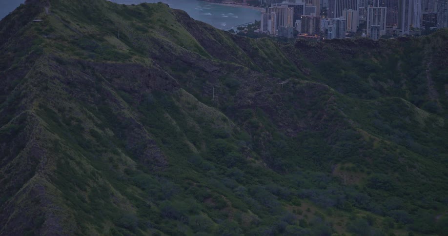 A slow aerial reveal shot tilts up from the lush green ridges of Diamond Head to the iconic Waikiki skyline at sunset
