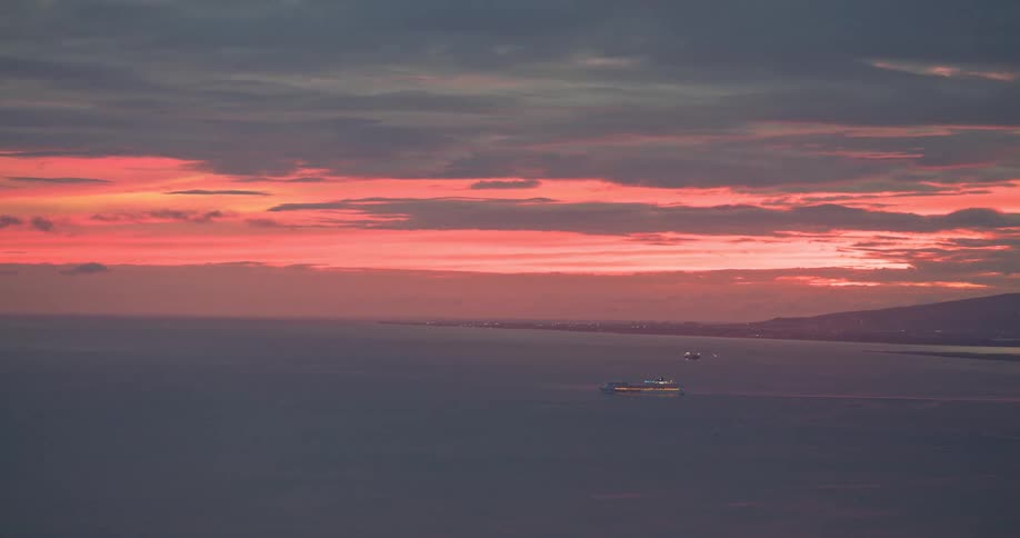 Cargo ships sail across the calm sea at dusk as a fiery red sunset paints the cloudy sky