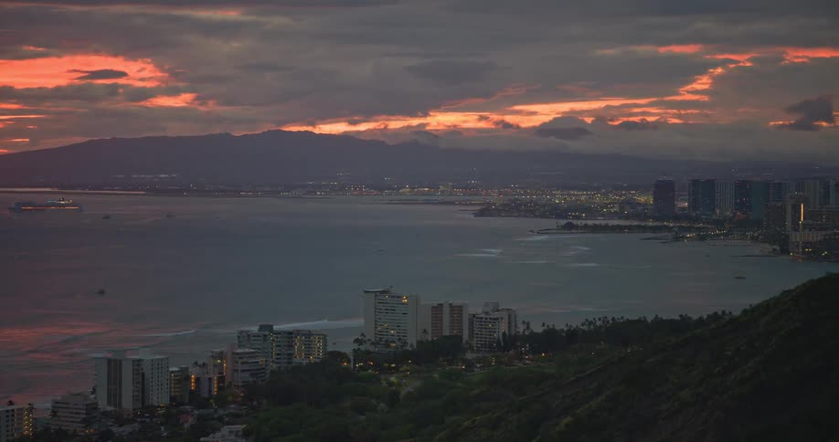 A dramatic, cloudy orange sunset glows over the coastal city of Honolulu, Hawaii, as city lights begin to twinkle