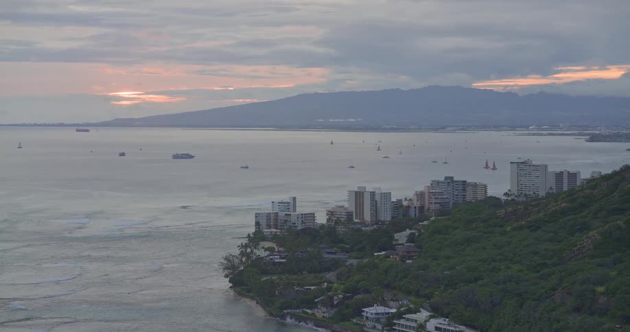 A scenic view of the Hawaiian coastline shows boats scattered across the ocean at sunset under a cloudy sky