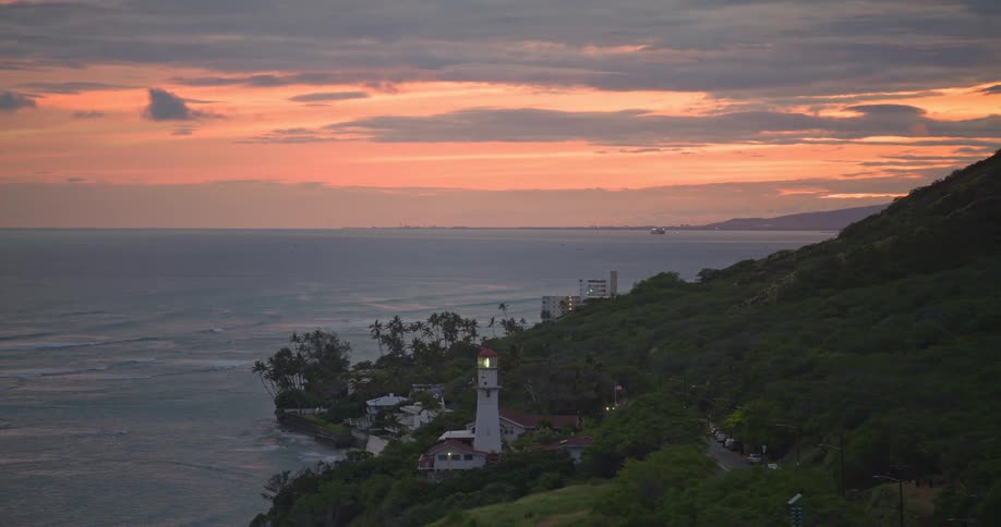 An aerial pull-back reveals the iconic Diamond Head Lighthouse on the Oahu, Hawaii coastline during a beautiful orange sunset