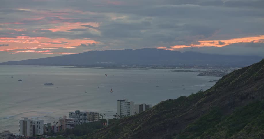 A beautiful panoramic view from a lush mountain reveals the vast ocean, a coastal city, and distant mountains under a cloudy sunset sky
