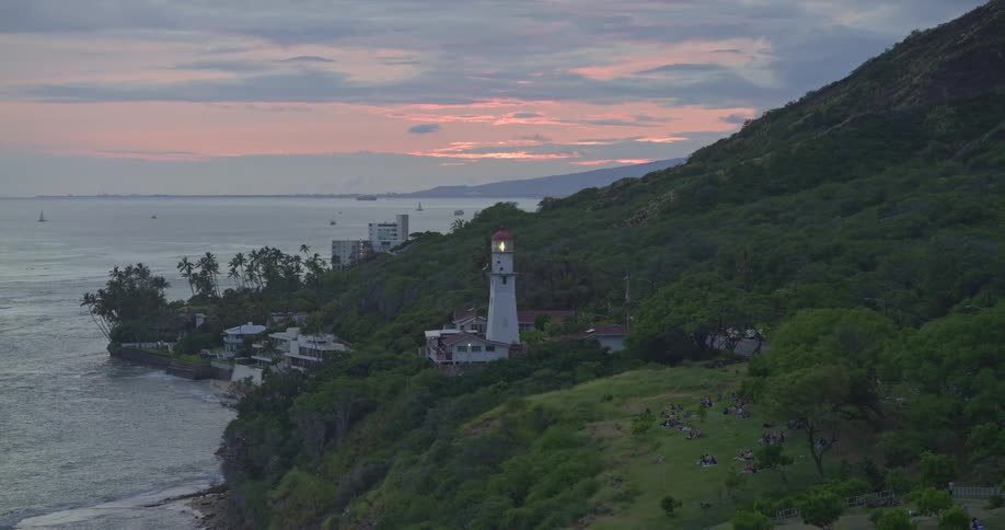 The Diamond Head Lighthouse in Hawaii shines its beacon over the Pacific Ocean at twilight as people relax on a nearby hill