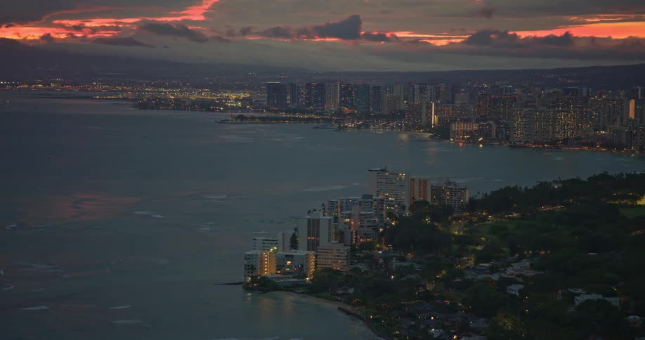 Twilight descends over the illuminated Honolulu coastline as a dramatic sunset paints the sky