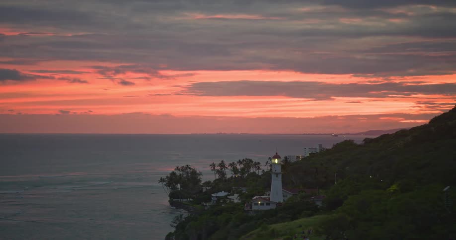 An aerial view of the Diamond Head Lighthouse on the coast of Oahu, Hawaii, as its beacon shines during a beautiful orange sunset