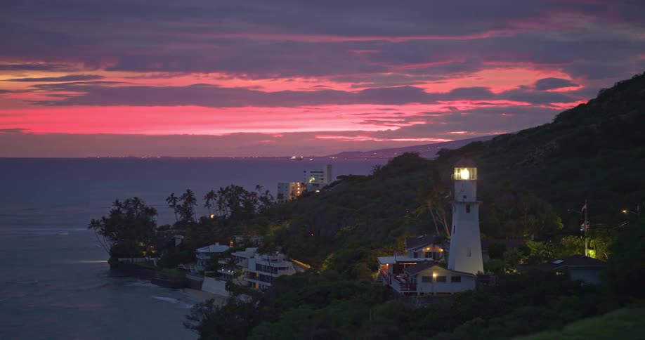 A coastal lighthouse illuminates a lush, tropical shoreline under a spectacular pink and purple sunset sky as the camera pulls back