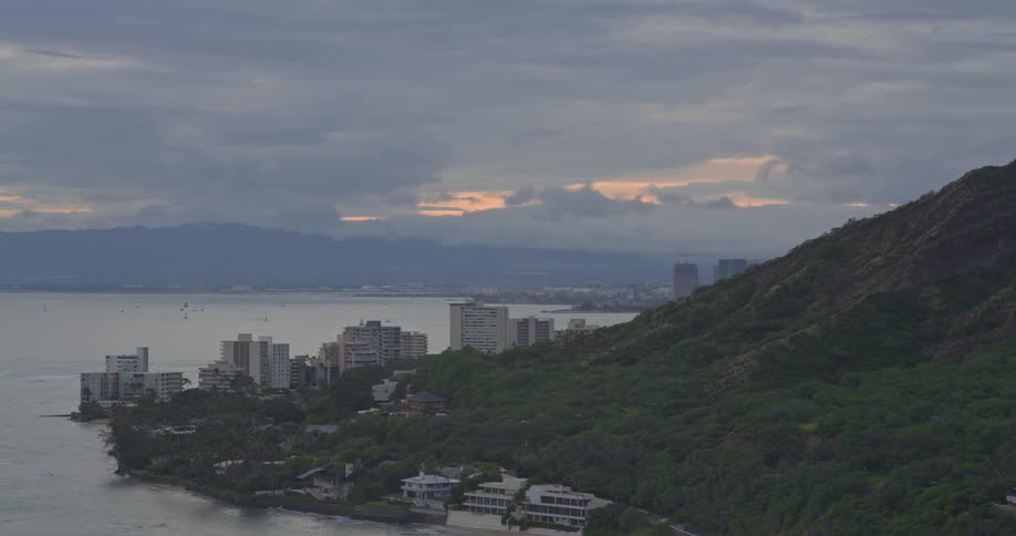 A wide aerial pan reveals the Honolulu coastline and Diamond Head mountain at dusk under a cloudy, dramatic sky