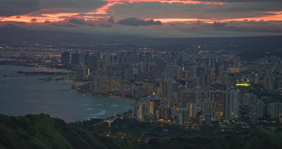 An expansive aerial view of Honolulu and Waikiki Beach reveals the city lights glowing at dusk under a dramatic and colorful sunset