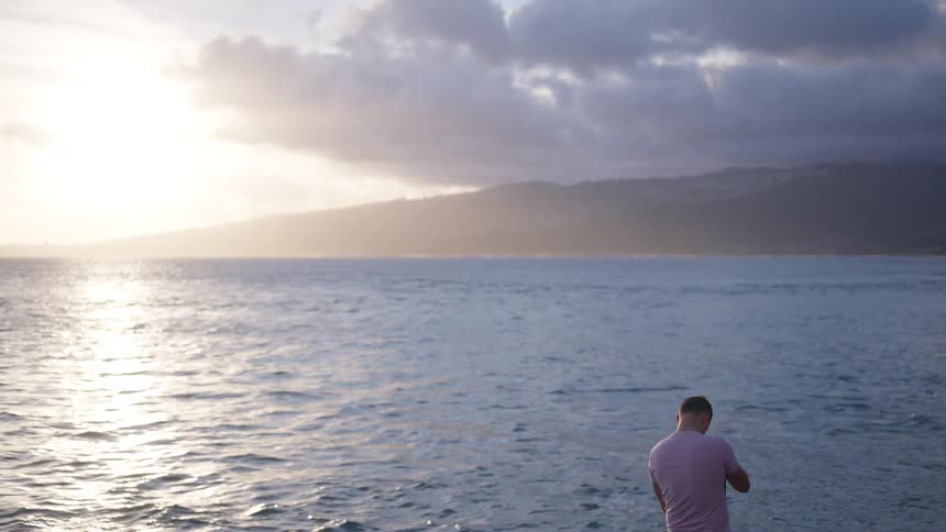 A male photographer stands on the shore, looking out over the tranquil ocean during a beautiful golden hour sunset