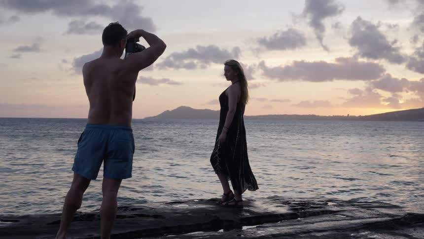 A male photographer takes pictures of a woman in a black dress posing on a rocky pier by the ocean during a scenic sunset