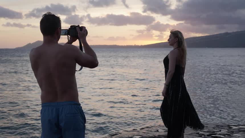 A man takes a photograph of a woman in a black dress as she poses by the ocean during a beautiful sunset