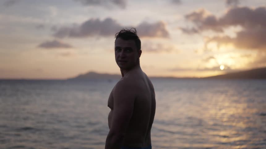 A shirtless man stands on the shore watching the ocean at sunset before turning to look thoughtfully over his shoulder