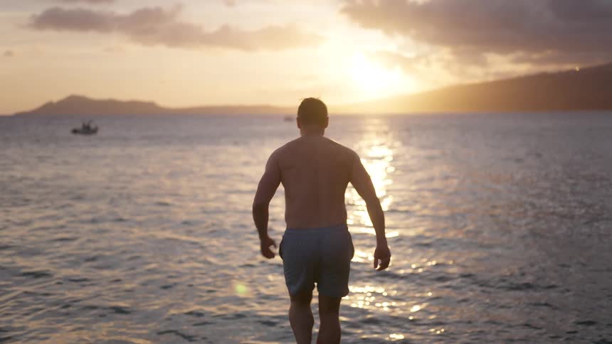 A shirtless man runs and joyfully jumps into the ocean from a rocky shore during a beautiful golden sunset, filmed in slow motion