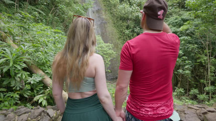 A romantic couple holds hands while walking towards a beautiful waterfall in a lush tropical jungle