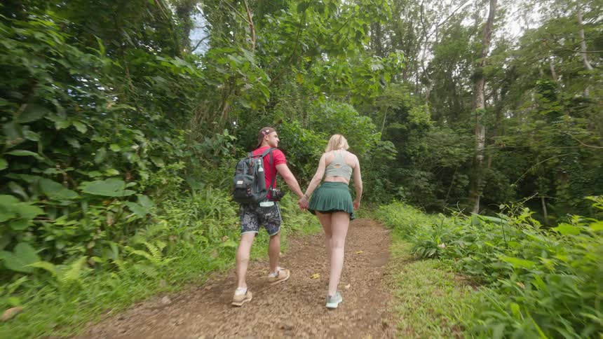 Slow motion rear view of an adventurous couple holding hands while hiking on a trail in a lush green jungle