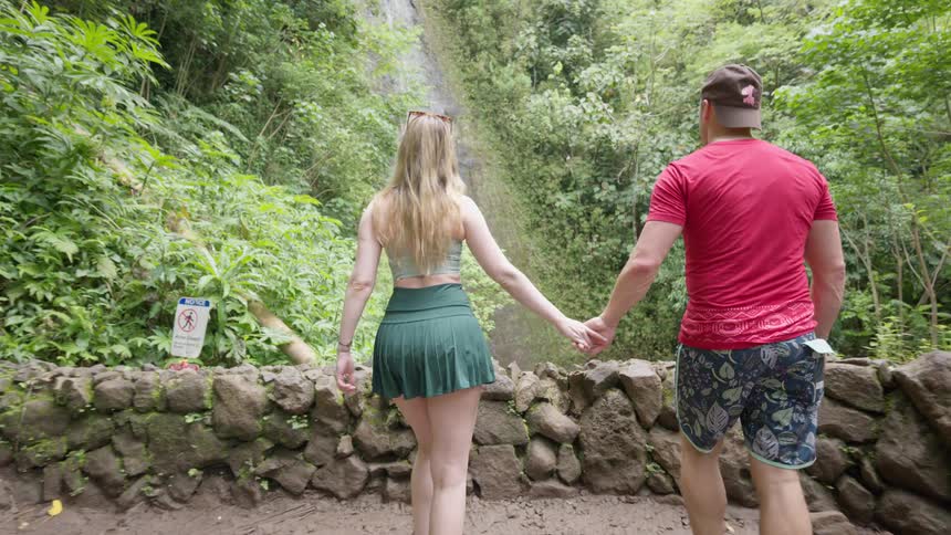 A young couple holds hands and walks together toward a scenic waterfall hidden deep within a lush tropical rainforest