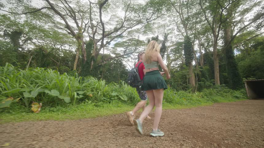 A young couple walks in slow motion down a path surrounded by a lush green jungle on an overcast day