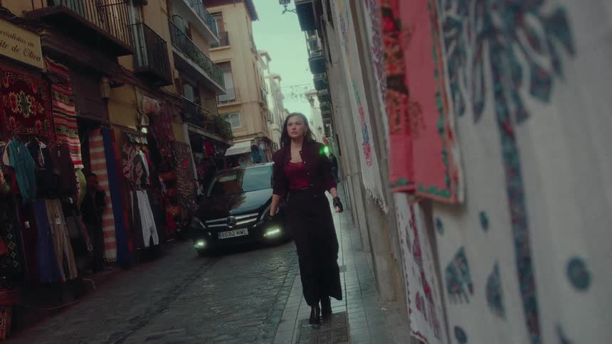 A fashionable woman walks with purpose in slow motion down a narrow, cobblestone European market street