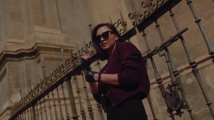A stylish woman in sunglasses and leather gloves leans against an old iron gate and looks at the camera