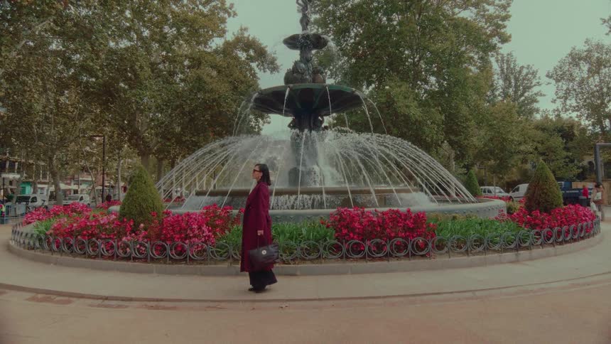 An elegant woman in a maroon coat stands pensively in a city park in front of an ornate water fountain