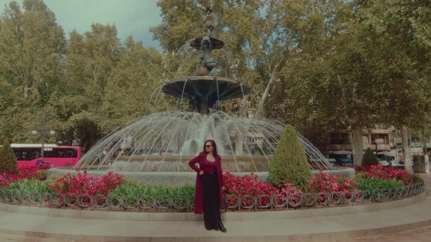 An elegant woman poses for a photo in front of a large, ornate fountain surrounded by flowers in a city park