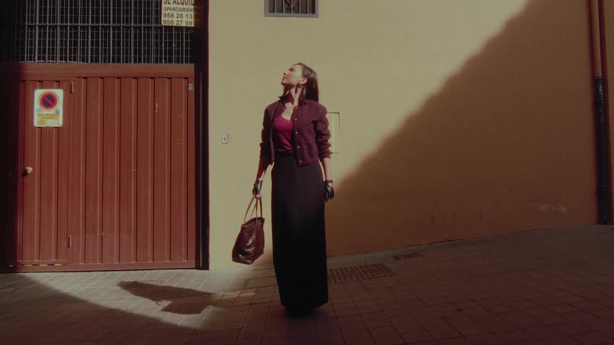 A stylish woman stands pensively in a sunlit city alley, looking up towards the sky