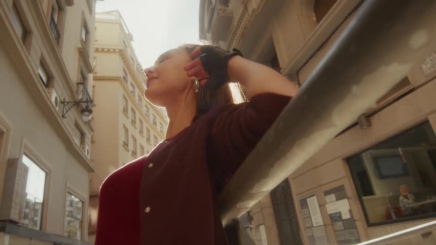 A stylish woman basks in the warm sunlight on a narrow European street, smiling serenely at the camera