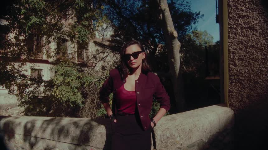 A stylish woman in sunglasses and a burgundy jacket poses confidently with her hands on her hips on a sun-dappled stone bridge