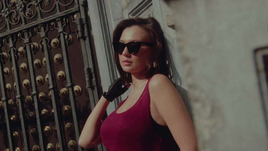 A mysterious and stylish woman in sunglasses and leather gloves poses confidently against a stone wall by an ornate gate in slow motion