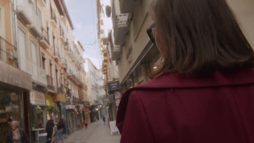 An over-the-shoulder slow-motion shot follows a stylish woman as she strolls down a bustling European city alley
