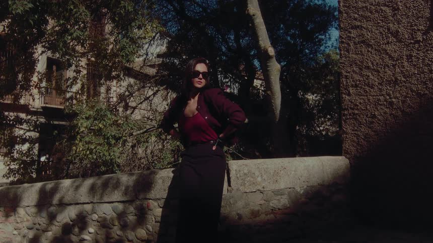 Confident woman in sunglasses and fashionable attire strikes a pose in a shadowy stone alleyway on a sunny day