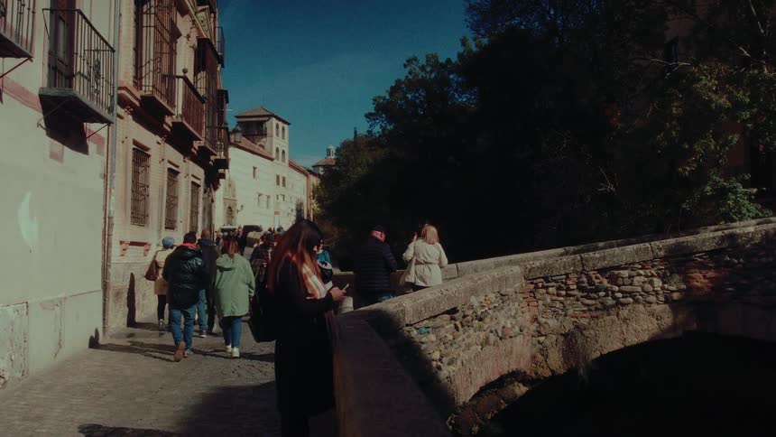 Tourists stroll along a historic cobblestone street next to an ancient stone bridge in a European city