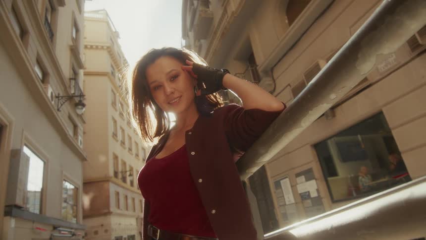 A confident and stylish woman smiles playfully for the camera, backlit by the bright sun on a beautiful city street