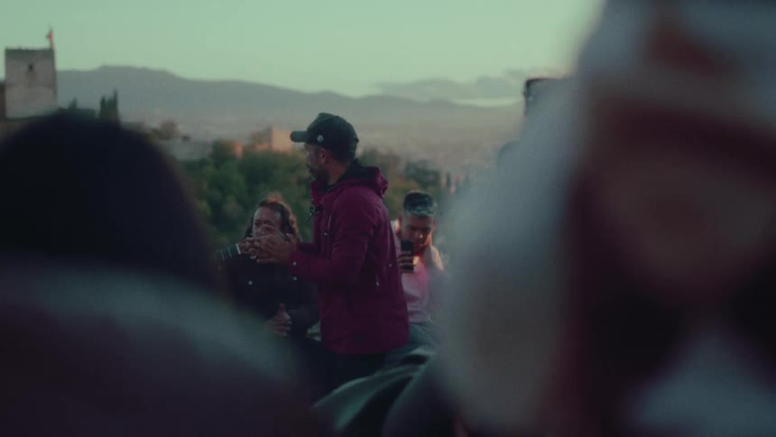 A group of people clap along to a guitar performance at a scenic viewpoint overlooking a historic city at sunset
