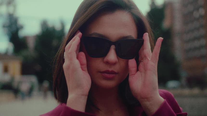 A stylish young woman pulls her sunglasses from her purse and confidently puts them on, looking directly into the camera in slow motion