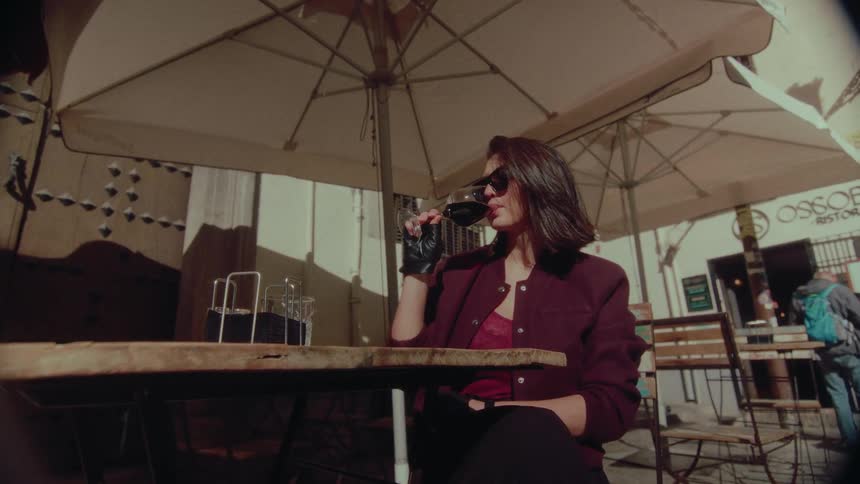 A chic woman in sunglasses and leather gloves slowly sips red wine at a sunny outdoor cafe