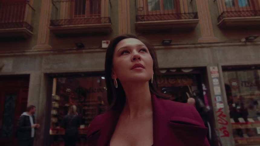 A low-angle cinematic shot of a stylish woman in a maroon coat looking up in awe while walking through a historic European city street
